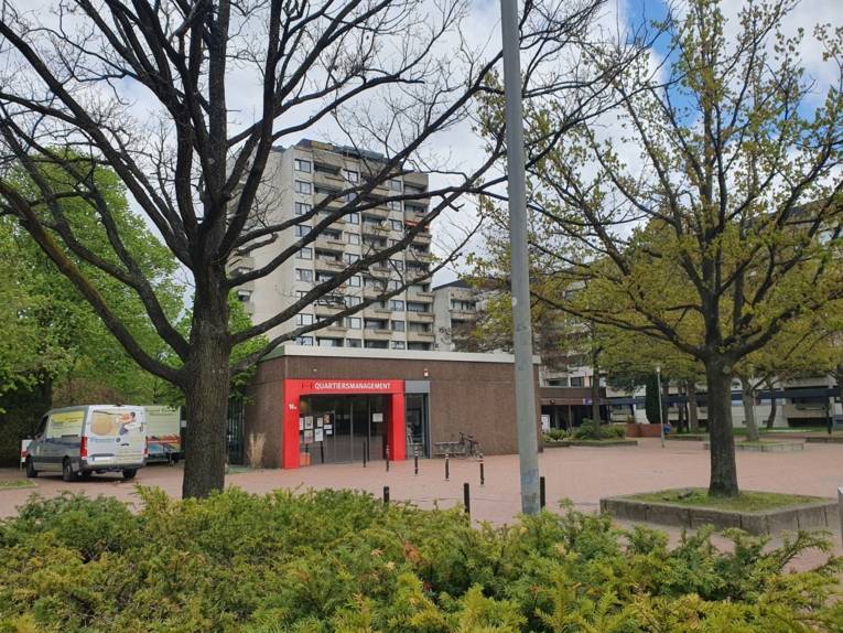 Im Vordergrund B&auml;ume, dahinter ein Platz mit einem Flachbau mit rotem Rahmen um den Eingang und der Aufschrift "Quartiersmanagement". Hinter dem Flachdachbau steht ein Hochhaus.