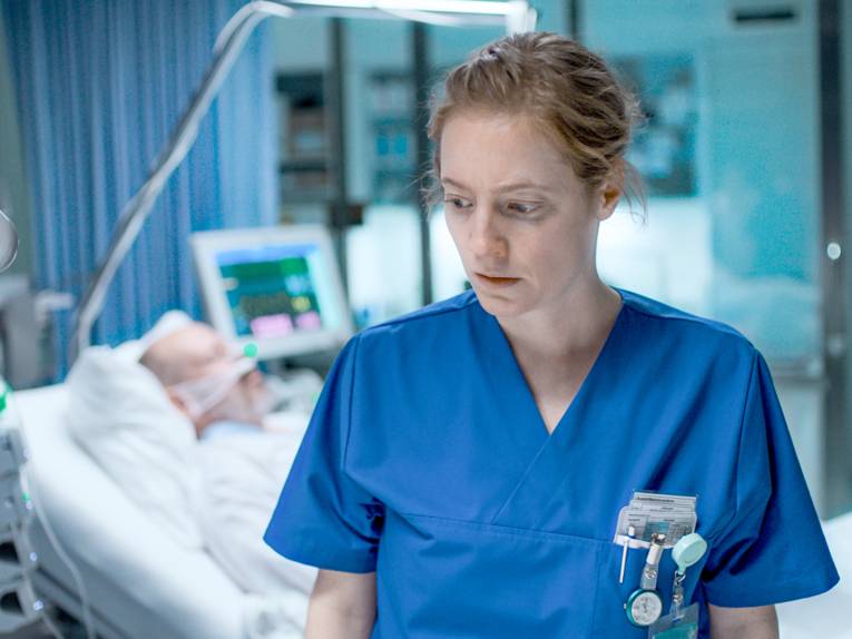 eine Frau in einer dunkelblauen Pfleger*innenkleidung. Im unscharfen Hintergrund liegt ein Mann in einem Krankenhausbett