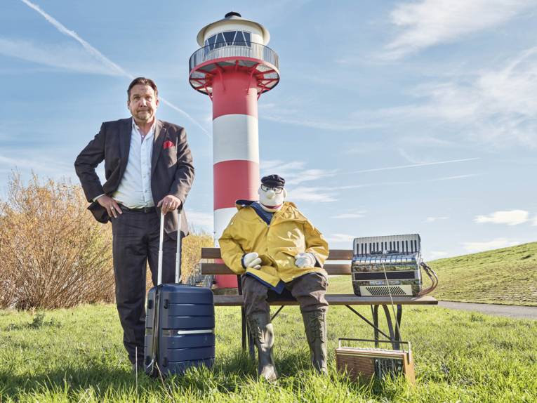 Auf einer Bank in einer Wiesenlandschaft mit Leuchtturm sitzt eine Puppe mit Friesennerz, Kappe und Akkordeon, daneben steht ein Mann im Anzug mit einem Rollkoffer.