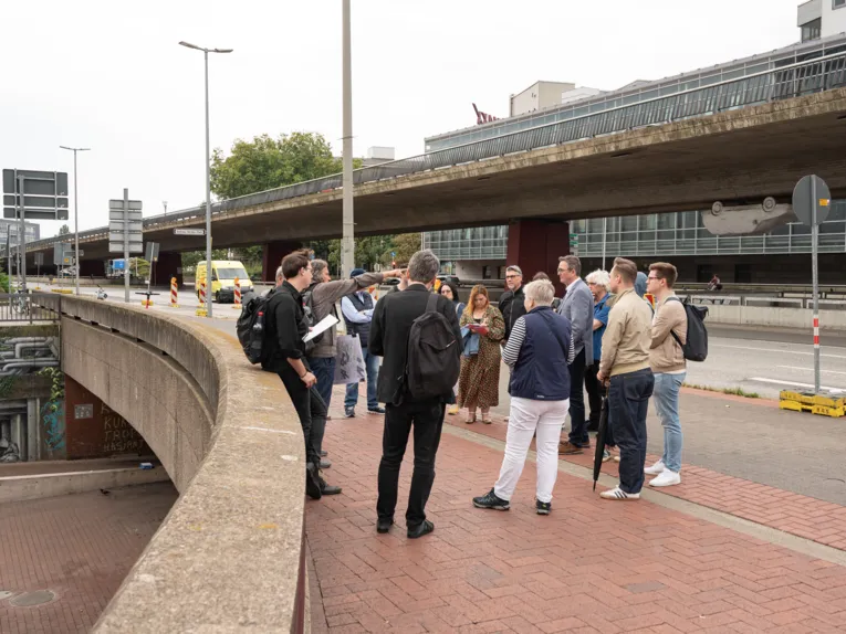 Eine Gruppe von Menschen steht am Rand einer Stra&szlig;e, &uuml;ber die eine Hochstra&szlig;e verl&auml;uft, unter ihnen ist links eine Vertiefungsebene zu sehen. Links zeigt eine Person nach rechts zur Stra&szlig;e.