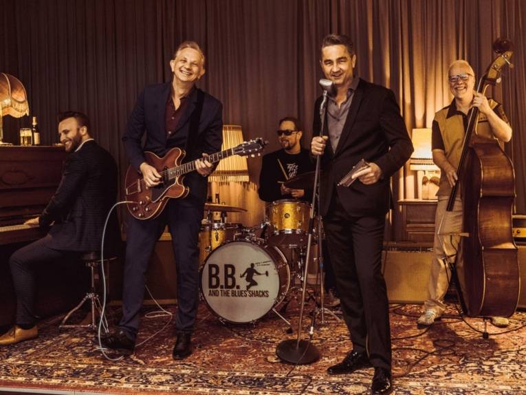 Fünf lachende Männer mit Gitarre, Kontrabass, Mikro, Piano und Schlagzeug in einem altmodischen Zimmer