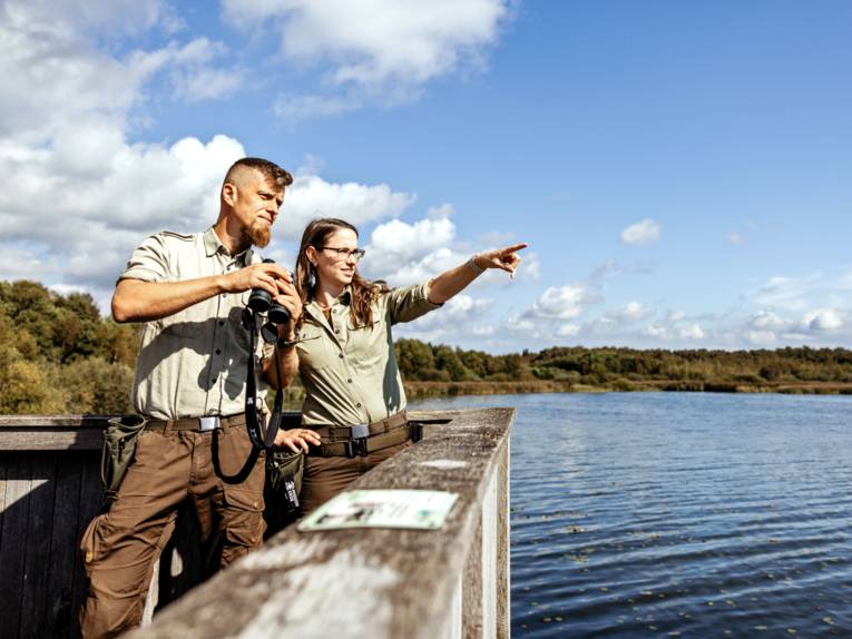 Eine Rangerin und ein Ranger stehen auf einer Aussichtsplattform. Er hält ein Fernglas in den Händen, sie zeigt mit dem Finger ein eine Richtung.