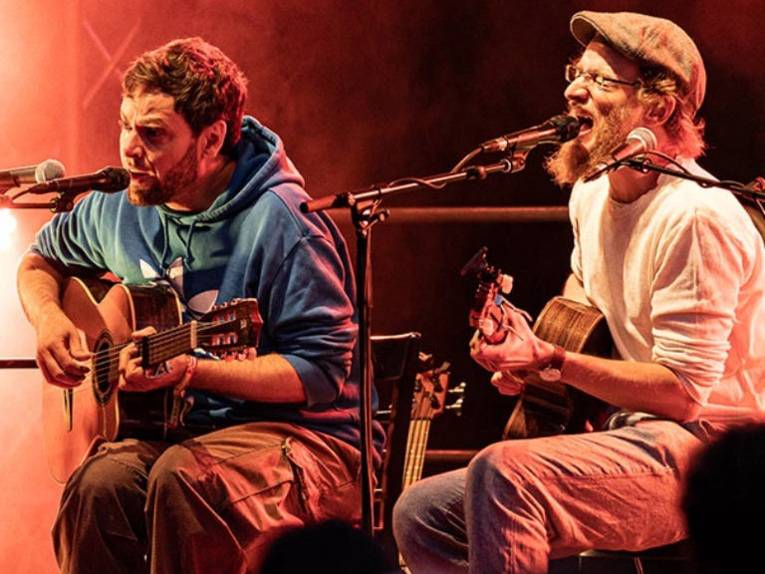 Zwei Männer sitzen mit Gitarren und Mikros auf einer Bühne.