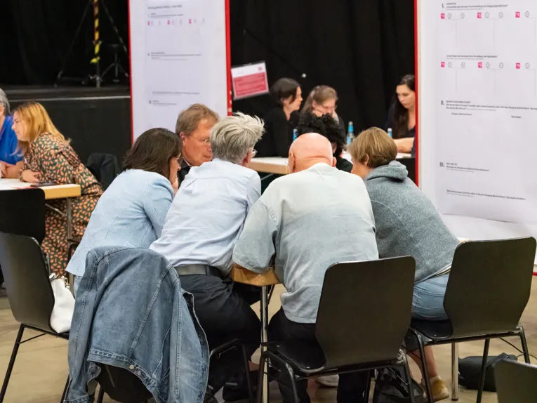 F&uuml;nf Menschen sitzen an einem Tisch und beugen sich zum Gespr&auml;ch nach vorn. Links steht eine Stellwand mit einem beschriebenen Papier. Hinter ihnen und links sind je eine weitere Gruppe an einem Tisch.