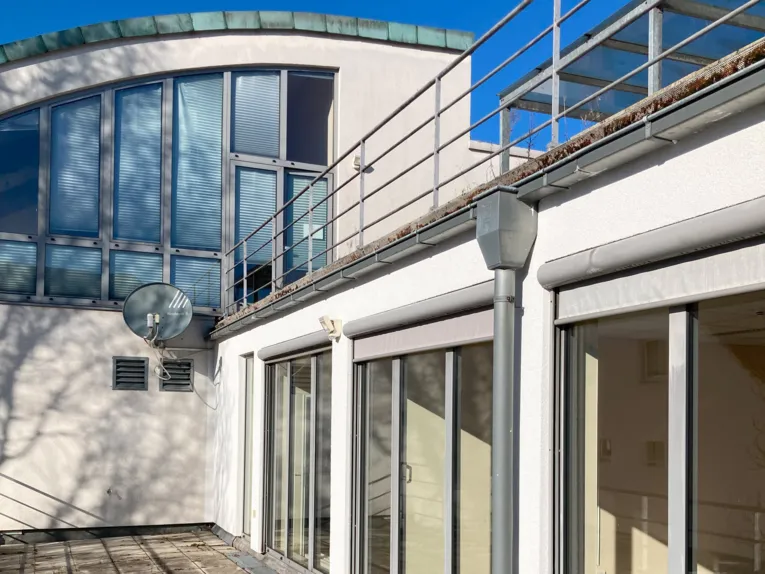 Ein weißer Gebäudeteil eines Dachs mit Dachterrasse vor hohen Fenstern, dahinter ein hoher Gebäudteil mit Fenstern und einem halbrunden Dach.