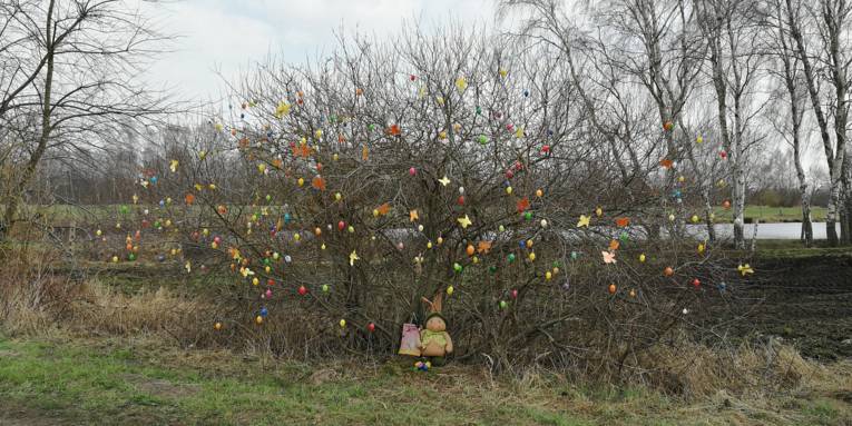 Bunte Eier und Schmetterlinge zieren einen Strauch. Am Stamm des Strauchs lehnt eine Osterhasenfigur.