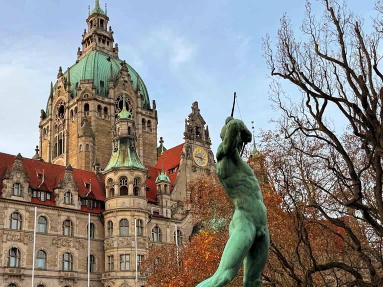 Blick aufs Neue Rathaus, im Vordergrund die Skulptur Bogenschütze, rechts ein Baum mit braunem Herbstlaub.