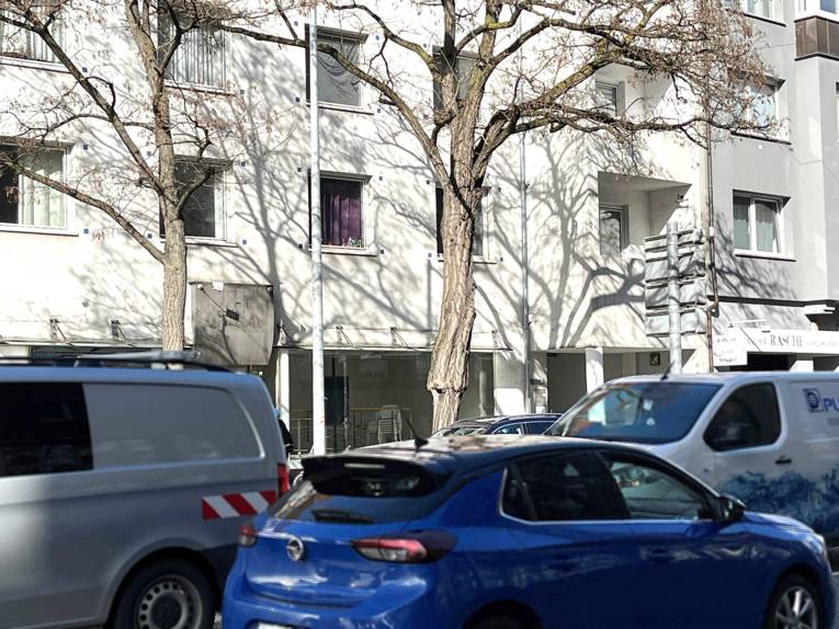 Drei Autos dicht aneinander im Verkehr auf einer Straße vor einem weißen Wohnhaus, dazwischen ein kahler Baum.