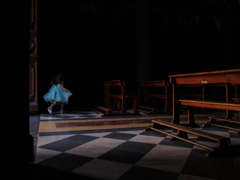 Zu sehen ist eine Fotografie von Simon Belperio: Ein Mädchen gekleidet in einem blauen Rüschenkledi läuft durch eine dunkle Kirche.