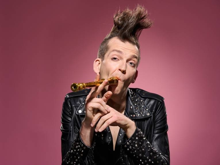 Zu sehen ist der Kabarettist Gabor Vosteen, er hat einen Iro, trägt eine Lederjacke und hält eine Flöte wie eine Zigarre am Mund.