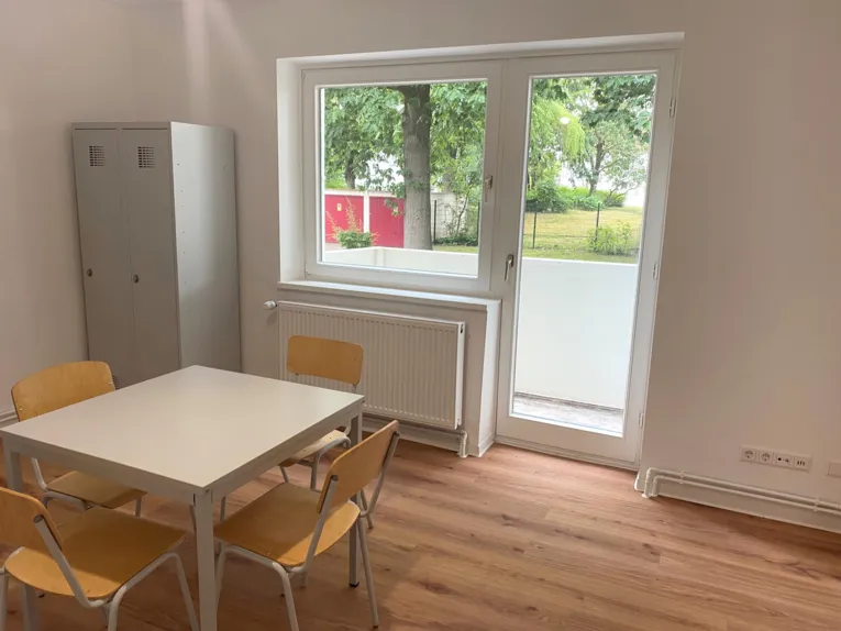 Ein Tisch mit vier St&uuml;hlen in einem Raum, dahinter ein Doppel-Spind, ein Fenster und eine Balkont&uuml;r zu einem kleinen Balkon.