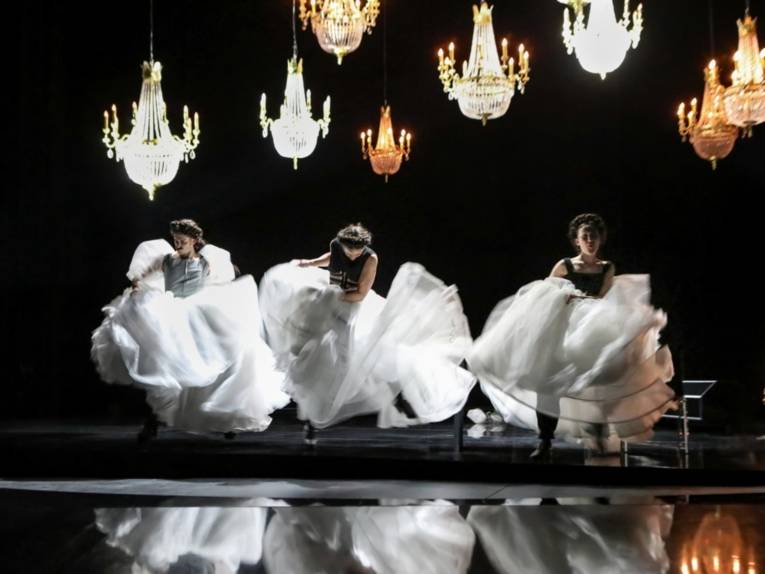 Drei Frauen tanzen wild in weißen Ballkleidern auf einer schwarz gehaltenen Bühne, bei der Kronleuchter von der Decke hängen.