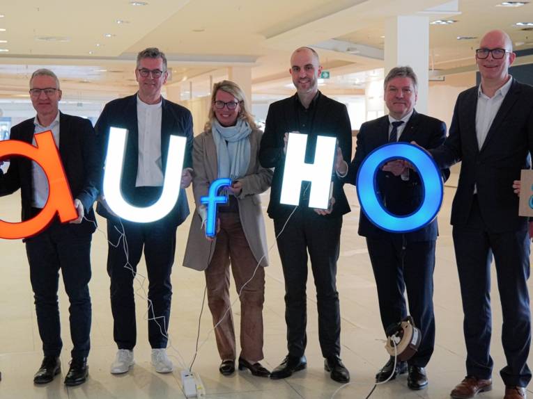 Acht Personen, die leuchtende Buchstaben halten. Diese bilden das Wort "aufhof".