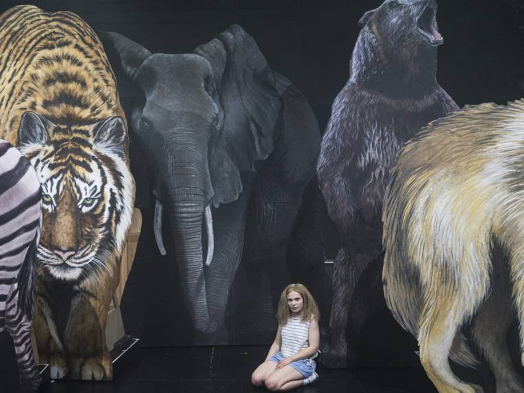 Eine junge Frau sitzt auf dem Boden einer Bühne, im Hintergrund sind überlebensgroße Bilder von wilden Tieren wie Elefanten und Tiger zu sehen.