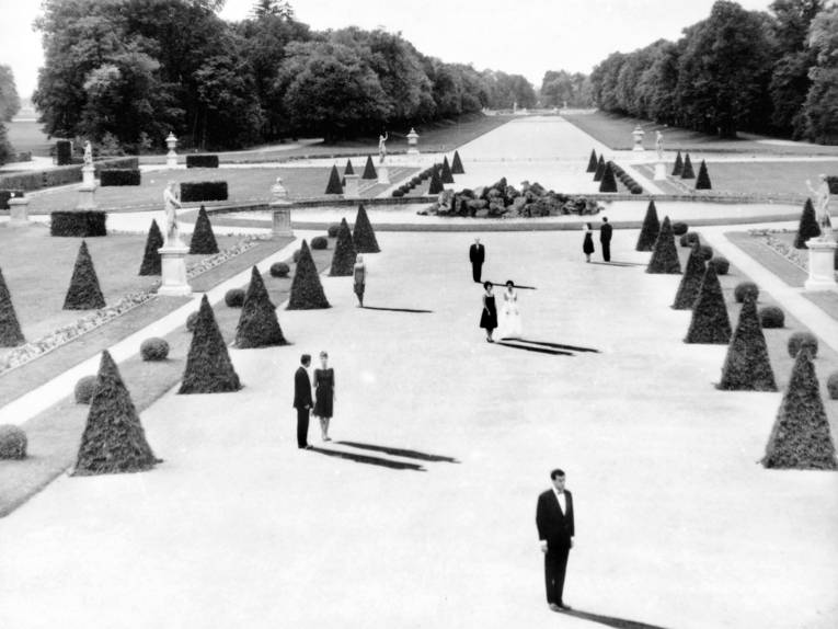 Ein breiter Weg in einem Park mit dreieckigen Büschen, einem großen Springbrunnen in der Mitte und Büsten. Die Personen werfen lange Schatten.