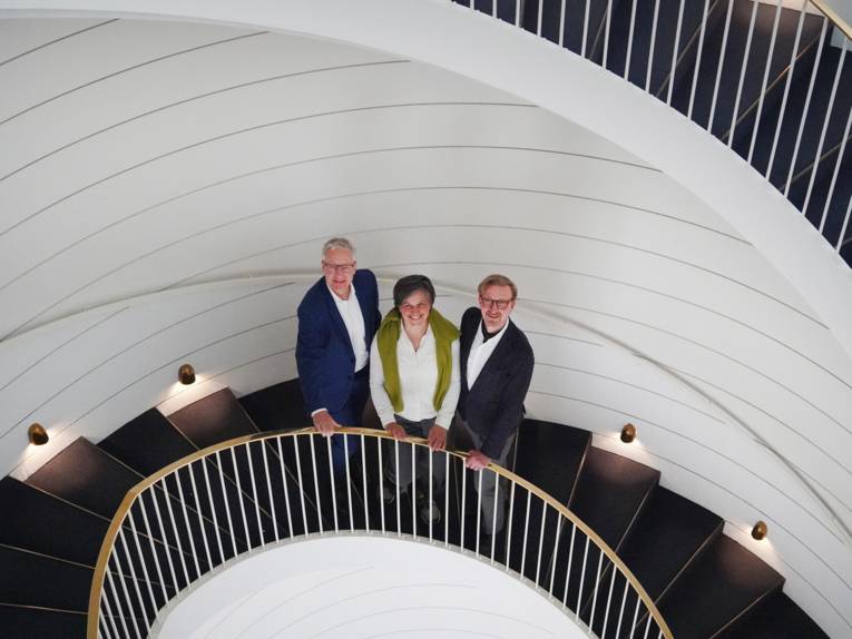 Blick von oben auf eine kreisrunde dunkle Treppe vor weißer Wand und mit weiß-goldenem Geländer, die sich wie ein Schneckenhaus windet. Auf der Treppe stehen eine Frau und zwei Männer.