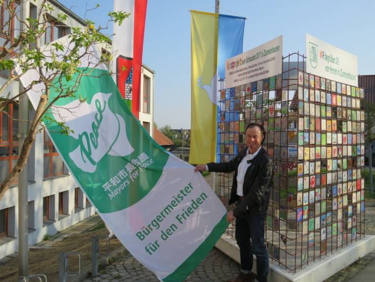 Ein Mann hält die Mayors-for-Peace-Flagge vor dem Rathaus in Zusmarshausen.