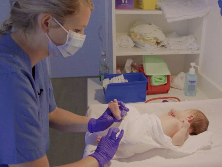 Eine Frau mit blauem Kittel, medizinischen Handschuhen und Atemschutzmaske wickelt einen Säugling.