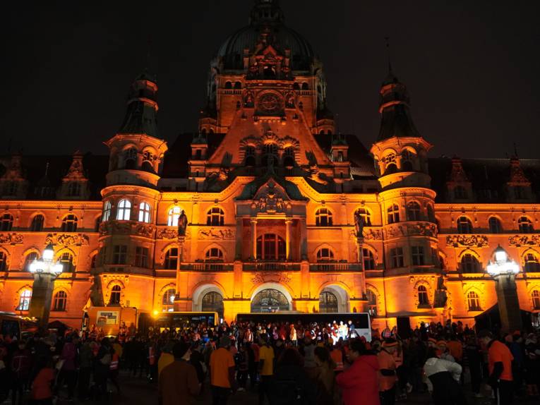 Ein altes schlossähnliches Gebäude im Dunkeln im orangem Licht angestrahlt. Davor stehen einige Menschen.