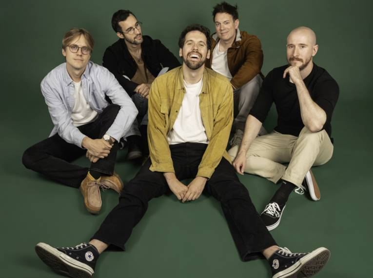 Lachender Mann sitzt mit ausgestreckten Beinen auf dem Boden, hinter ihm vier weitere Männer.