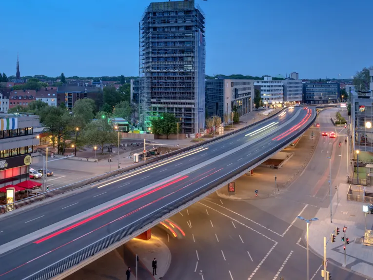 Eine Hochstraße von oben, darauf lang gezogene Lichter. Unter der Hochstraße eine Straße, im Hintergrund Gebäude.