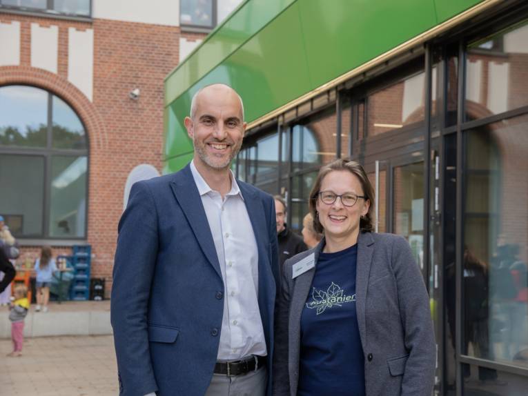 Ein Mann und eine Frau stehen vor einem Gebäude, links gemauert mit rundem Bogenfenster, rechts mit Glasfront und grünem Abschluss oben.