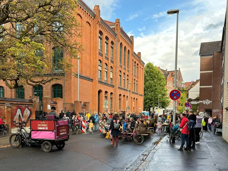 Auf einer Straße stehen und sitzen viele Menschen auf Bänken und an Tischen, die sich unterhalten.