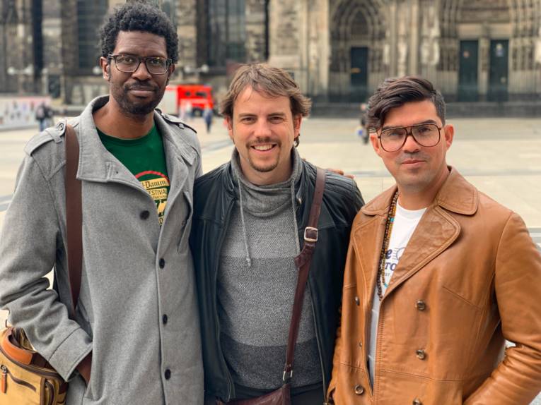 Drei Männer stehen nebeneinander auf der Domplatte vor dem Kölner Dom.