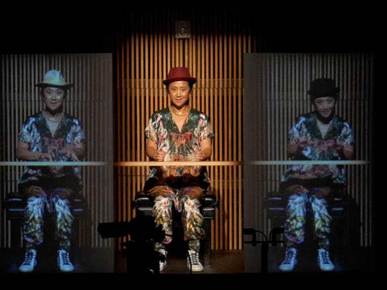 Eine Frau sitzt auf einer Bühne und spielt ein imaginäres Klavier, links und rechts von ihr sind lebensgroße Videoproduktion von ihr selbst.