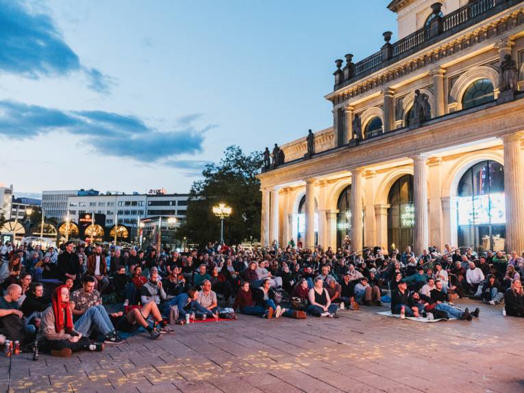 Auf dem Bild sitzen viele Menschen nebeinander auf dem Boden und rechts neben Ihnen ist die Staatsoper.