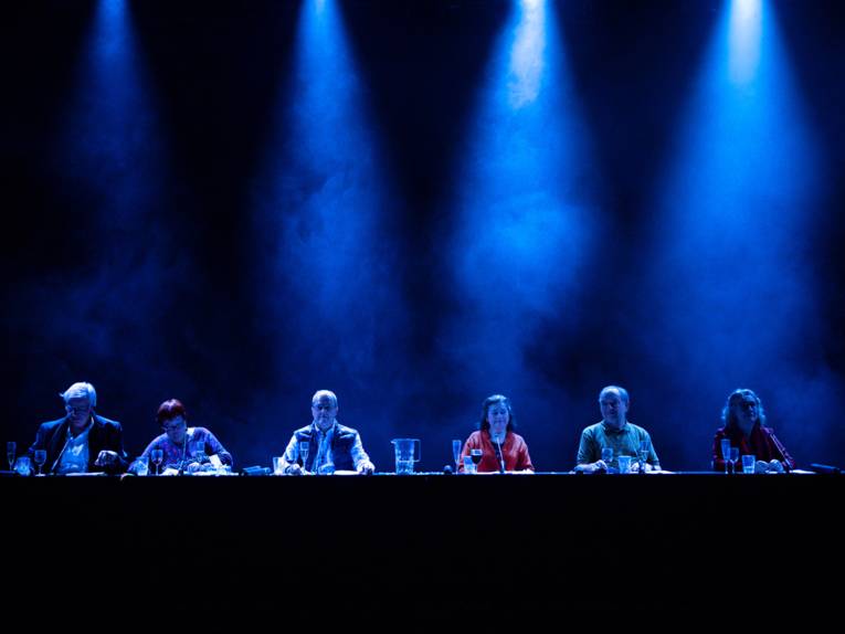 Drei Frauen und drei Männer sitzen auf einer Bühne nebeneinander an eine langen Tafel und werden von oben mit Kegelscheinwerfern beleuchtet.