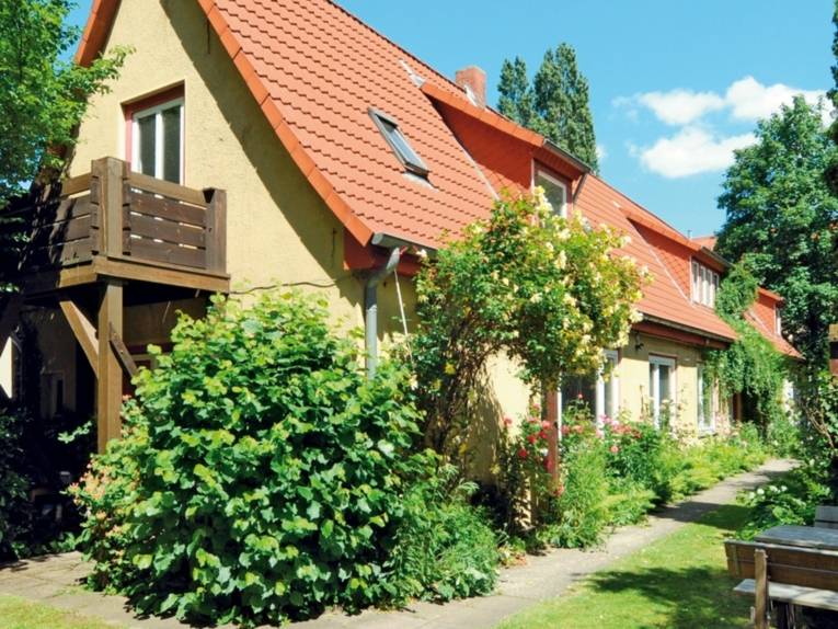 Haus mit Holzbalkon, umgeben von Bäumen und Büschen.