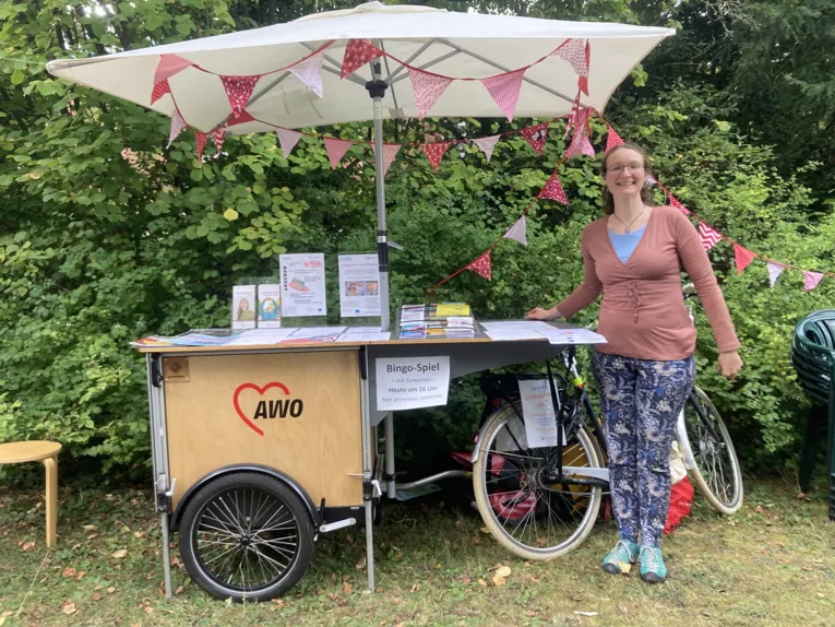 Ein Stand mit Wimpelketten und Schirm des AWo Seniorenb&uuml;ros und eine Frau l&auml;chelt