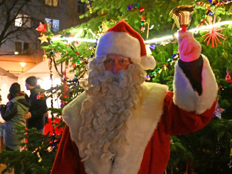 Auf dem Bild ist ein Weihnachtsmann zu sehen, der eine Glocke läutet. Im Hintergrund ist ein Weihnachtsmarkt im Dunkeln zu sehen.
