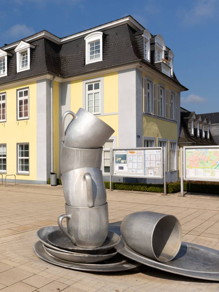 Gelbe Villa mit grüner Eingangstür und gepflastertem Fußgängerbereich. Davor ist ein Kunstwerk, das aussieht, wie ein überdimensionaler Stapel aus Geschirr.