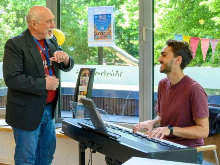 Ein Mann sitzt an einem elektrischen Tasteninstrument, vor ihm steht ein Mann, der etwas &auml;lter ist als er. Beide lachen.