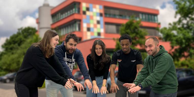 Fünd junge Menschen strecken vor dem Gebäude des Diakonie-Kolleg Hannover die Hände in Hüfthöhe voreinander aus und bilden dabei einen Viertelkreis.