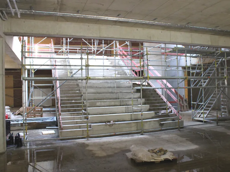 Ein Beton-Rohbau, in der Mitte eines gro&szlig;en Raums eine breite Treppe umgeben von Bauger&uuml;sten.