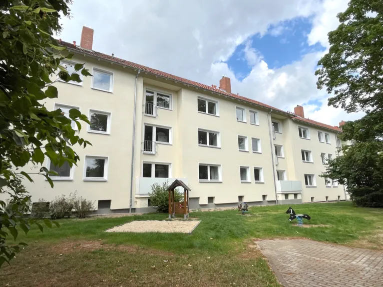 Dreigeschossige Wohnh&auml;user mit gelber Fassade, davor eine gr&uuml;ne Rasenfl&auml;che mit einem Sandkasten, einem Spielh&auml;uschen und zwei Wipppferdchen.