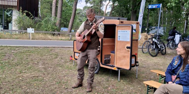 Ein Mann tr&auml;gt Outdoor-Kleidung, steht vor einem besonderen Fahrradanh&auml;nger aus Holz mit Mikrofontechnik und Infomaterial, er spielt auf einer Gitarre.