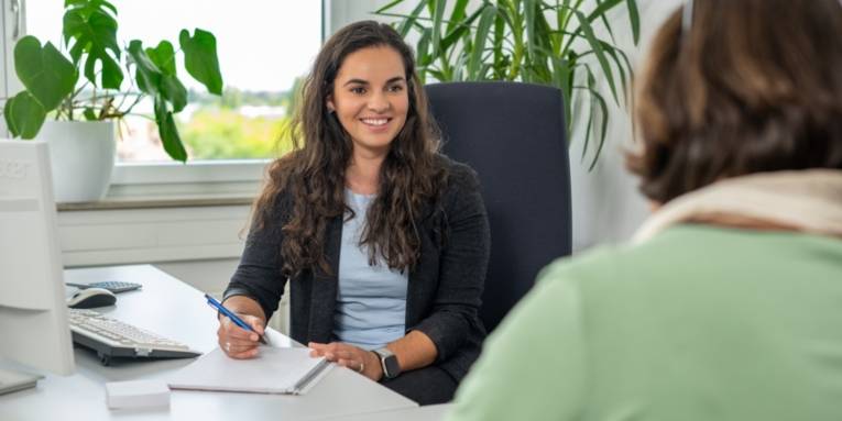 Zwei Personen sitzen sich an einem Büroschreibtisch gegenüber und sprechen miteinander