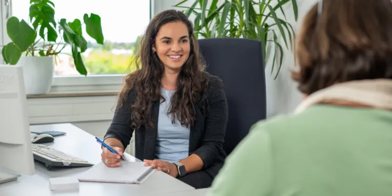 Zwei Personen sitzen sich an einem Büroschreibtisch gegenüber und sprechen miteinander