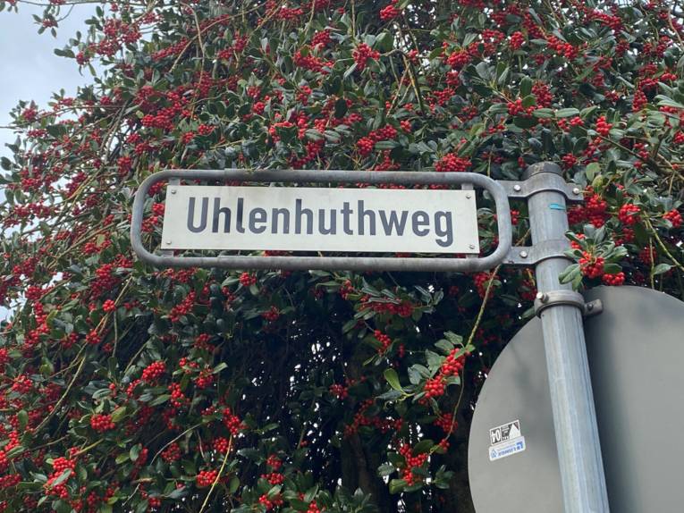 Auf dem Bild ist ein Straßenschild zu sehen mit dem Namen "Uhlenhuthweg". Hinter dem Straßenschild ist ein Baum mit roten Beeren zu sehen.