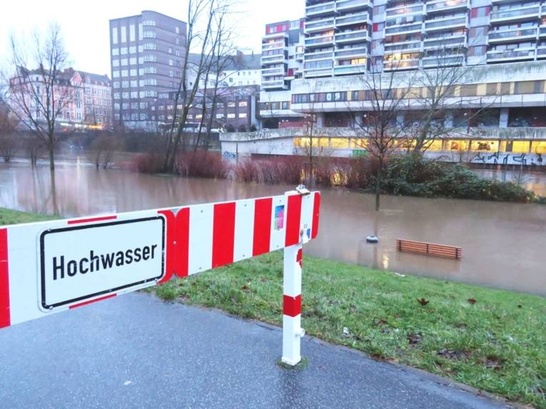 Ein rot-weißes Sperrschild mit der Aufschrift Hochwasser, dahinter eine überflutete Wiese, ein voller Fluss und ein Hochhaus.