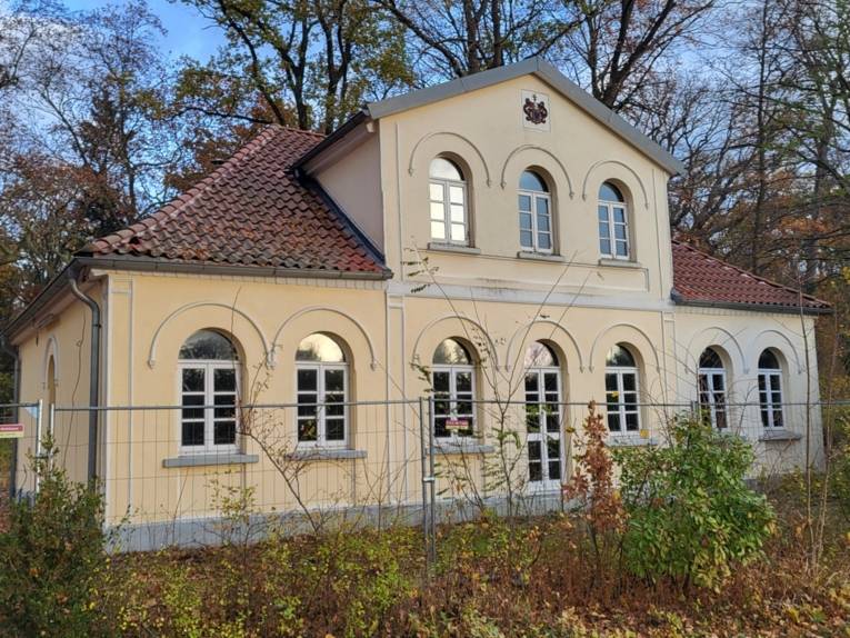 Ein helles beiges Gebäude umgeben von Bäumen mit einem Obergeschoss, unten in der Front sechs oben abgerundete Fenster mit Butzenscheiben, oben im Giebel drei ebensolche Fenster.