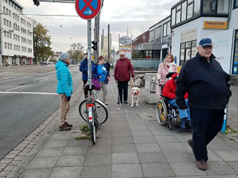Menschen und Menschen mit Behinderung (Blinde Personen, eine Person mit Blindenhund und eine Person im Rollstuhl) auf der Sutelstra&szlig;e.