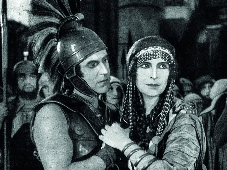 Eine historische Filmszene in Schwarz-Weiß: Zwei Personen in aufwendig gestalteten antiken Kostümen stehen eng beieinander. Eine Person trägt eine Rüstung mit Helm und Federbusch, die andere ein reich verziertes Gewand mit Kopfschmuck. Im Hintergrund sind weitere Personen sowie eine monumentale Innenkulisse zu erkennen. Die Aufnahme erinnert an einen Stummfilm oder ein frühes Historienepos.