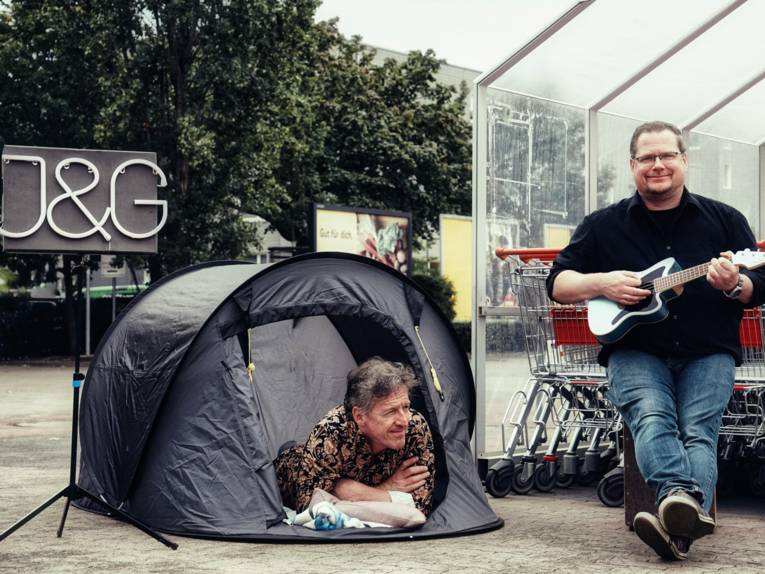 Zwei Männer auf einem Supermarktparkplatz, der eine liegt bäuchlings in einem kleinen Zelt, der andere sitzt mit Gitarre vor einem Unterstand mit Einkaufswagen.