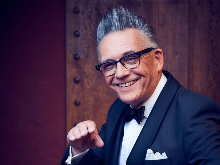 Zu sehen ist ein Mann mit grauen aufgestellten Haaren und einem Anzug vor einer Holz vertäfelten Wand.