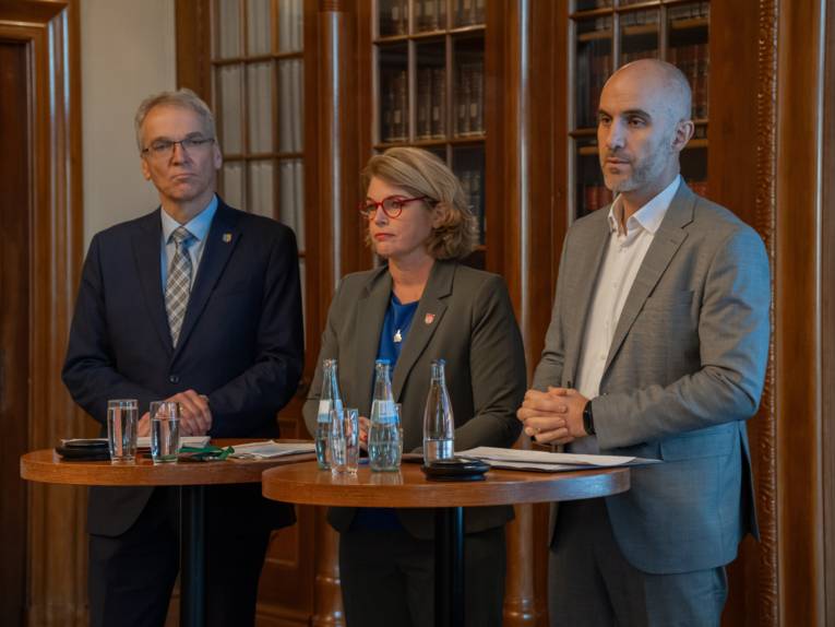 Drei Personen stehen an Stehtischen, vor ihnen stehen Wassergläser.